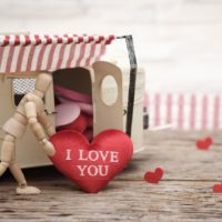 Wooden mannequin figure pushing a red heart labeled 'I LOVE YOU' towards a toy camper, illustrating emotional expression in relationships