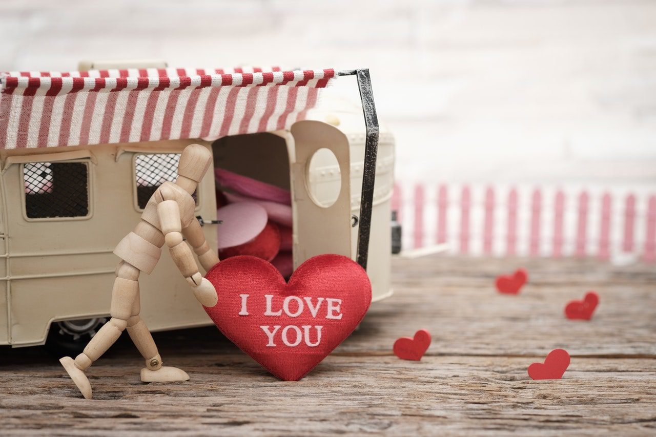 Wooden mannequin figure pushing a red heart labeled 'I LOVE YOU' towards a toy camper, illustrating emotional expression in relationships