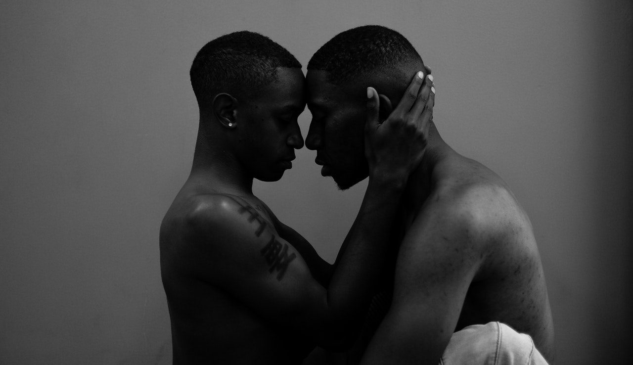 Black and white portrait of two shirtless men standing profile to profile, holding head to head with hands on each other's faces