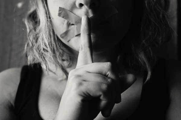 Close-up of a person with tape over their mouth holding a finger up to quiet disputes.