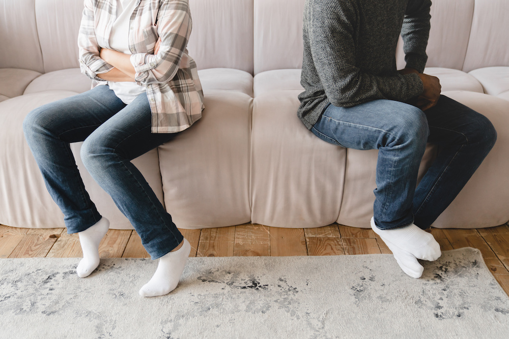 A couple sitting on opposite ends of a sofa with arms crossed during a conflict. A couple sitting on opposite ends of a sofa with arms crossed during a conflict.