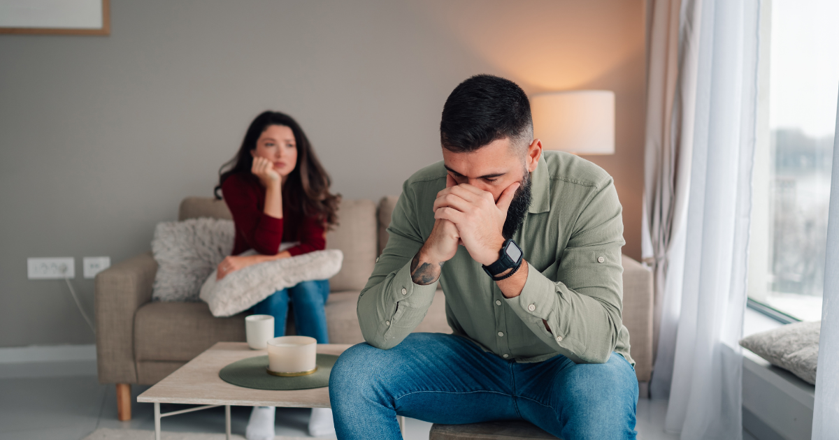 Stressed man sitting in the foreground holding his face in his hands while a distant woman sits thoughtfully on the sofa behind him