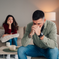 Stressed man sitting in the foreground holding his face in his hands while a distant woman sits thoughtfully on the sofa behind him