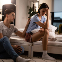 Young man sitting on the floor reaching up to place a hand on the back of his sad partner who is sitting on the bed holding her head