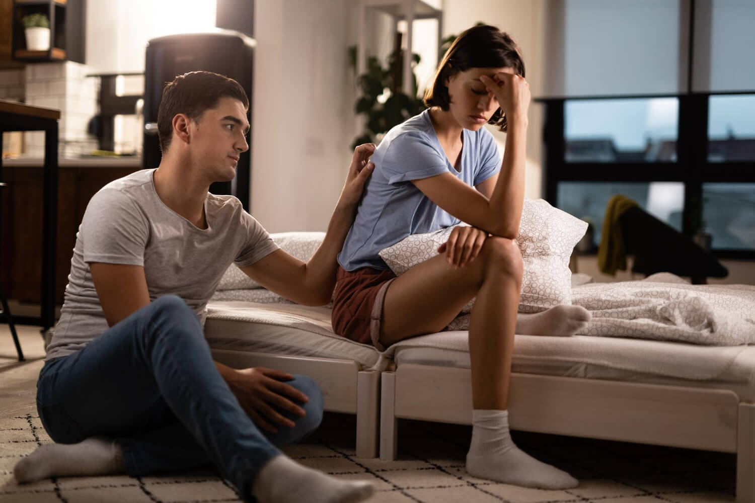 Young man sitting on the floor reaching up to place a hand on the back of his sad partner who is sitting on the bed holding her head